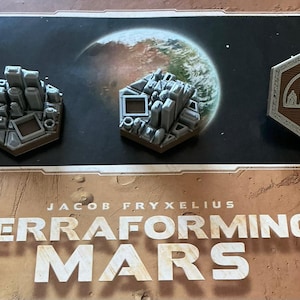 Peut inclure: Trois pièces du jeu de société Terraforming Mars. Deux jetons de ville gris et un jeton de ville marron avec un motif de paysage urbain sont affichés sur un plateau de jeu avec le titre du jeu.