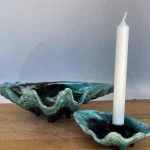 May include: A large, deep, shell-shaped ceramic bowl with a textured, wavy rim in shades of dark teal and green, accompanied by a smaller matching shell-shaped candle holder containing a single, tall, slender white taper candle.