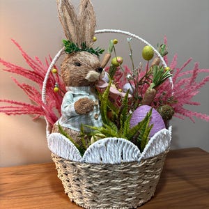 Könnte beinhalten: Osterkorb mit einem Strohkaninchen in einem grün-weiß karierten Hemd. Der Korb ist mit Kunstkarotten, Eiern und rosa und grünen Zierpflanzen gefüllt. Der Korb hat eine weiße, gewellte Bordüre und einen weißen Griff.