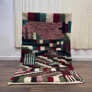 Peut inclure: Un tapis rectangulaire fait à la main avec un motif géométrique. Le tapis présente un mélange de couleurs, dont le bordeaux, le vert, le noir et le crème. Le design intègre des carrés, des rectangles et des rayures, créant un aspect texturé. Le tapis est posé sur un sol en bois.