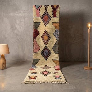 Peut inclure: Un long tapis de couloir rectangulaire à fond crème orné d'un motif répétitif de losanges et de formes géométriques dans des tons de rose, rouge, bleu, jaune et vert foncé. Le tapis présente un poil épais et texturé avec des franges.