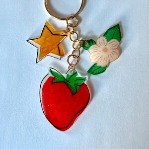 Puede incluir: Un llavero dorado con un dije de estrella, flor y fresa. La estrella es amarilla con un contorno blanco. La flor es rosa claro con hojas verdes. La fresa es roja con un tallo verde.