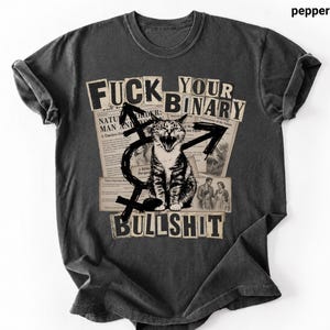 May include: Pepper-colored t-shirt with a distressed graphic featuring a cat, newspaper clippings, and the words "FUCK YOUR BINARY BULLSHIT." The design includes gender symbols and a vintage aesthetic.