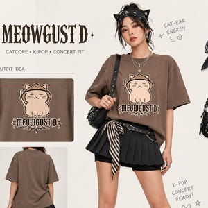 Może przedstawiać: Brązowa koszulka z grafiką kota i napisem "MEOWGUST D." Na zdjęciu widoczna jest r&oacute;wnież czarna plisowana sp&oacute;dnica, czarne buty i akcesoria, takie jak torebka, pierścionki i szalik. Widoczny jest r&oacute;wnież tekst "SOFT TEE. BIG ENERGY".