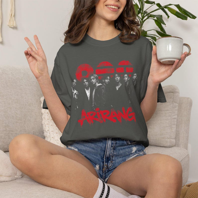 May include: A charcoal gray short-sleeve t-shirt featuring a black and white graphic of a group of seven individuals. Above the group are three red circular shapes, and below the group is the red text Arirang in a stylized, bold font.