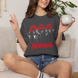 May include: A charcoal gray short-sleeve t-shirt featuring a black and white photographic print of a group of seven individuals. Above the group are three red circular shapes, and below is the red text ARIRANG in a stylized, dripping font.