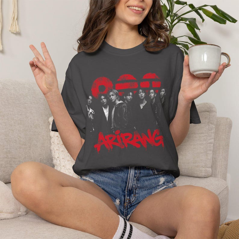May include: A charcoal gray short-sleeve t-shirt featuring a black and white photographic print of a group of seven individuals. Above the group are three red circular shapes, and below is the red text ARIRANG in a stylized, dripping font.