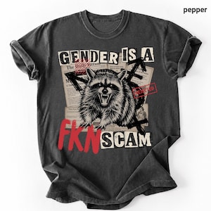 Puede incluir: Una camiseta gris carbón con un estampado gráfico de un mapache gruñendo sobre un collage de recortes de periódico. El texto dice GENDER IS A FKN SCAM en letras negras y rojas, con un sello rojo de REJECTED y varios símbolos negros.