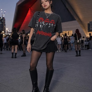 May include: A charcoal gray short-sleeve t-shirt featuring a graphic print of a group of people and the red text ARIRANG, paired with a black pleated mini skirt, sheer black tights, and black knee-high platform boots with a chunky sole.