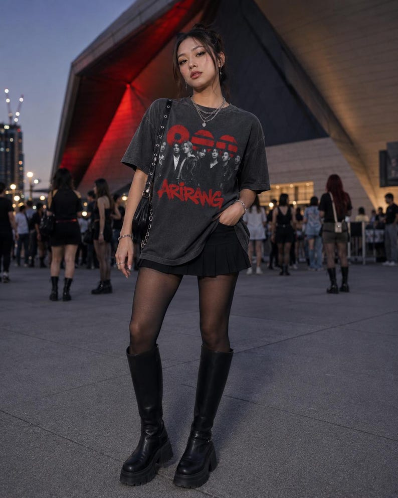 May include: A charcoal gray short-sleeve t-shirt featuring a graphic print of a group of people and the red text ARIRANG, paired with a black pleated mini skirt, sheer black tights, and black knee-high platform boots with a chunky sole.