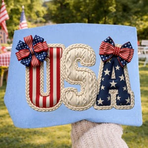 May include: A light blue sign with the word "USA" in a patriotic design. The letters are decorated with red, white, and blue accents, including bows and star patterns, perfect for celebrating the 4th of July.