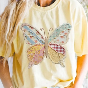 Puede incluir: Camiseta amarilla clara con un gran diseño de mariposa de patchwork. Las alas de la mariposa están compuestas por varias telas estampadas en tonos azules, rosas, rojos y amarillos. La camiseta tiene mangas cortas y cuello redondo. La mariposa mide unos 20 cm de ancho.