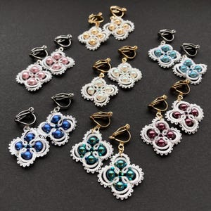 Può includere: Una collezione di orecchini a clip con pizzo chiacchierino bianco a forma di quadrifoglio, ciascuno con una perla centrale in vari colori tra cui rosa, oro, bianco, blu, verde acqua e viola scuro, dotati di clip in metallo.