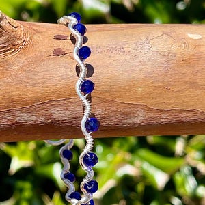 Może przedstawiać: Ręcznie robiony srebrny pierścionek z falistym wzorem, ozdobiony żywymi niebieskimi koralikami. Pierścionek jest prezentowany na naturalnym drewnianym tle z rozmytym zielonym tłem liści, prezentując delikatne rzemiosło.