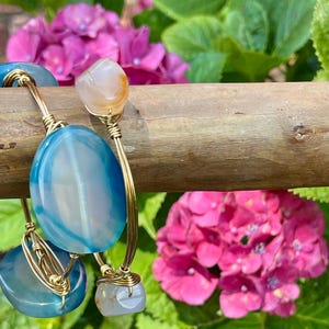 May include: A wire-wrapped bracelet featuring three polished blue agate stones and two smaller translucent white stones. The stones are secured with gold-toned wire, creating a bypass-style design that wraps around the wrist with open ends.