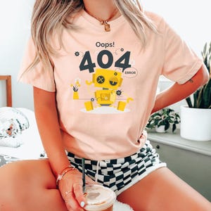May include: A light peach-colored t-shirt with a graphic of a yellow robot with the text "Oops! 404 Error". The robot has a square head and is sitting down. The person is wearing black and white checkered shorts.