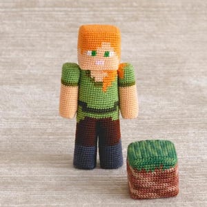 May include: A crochet doll featuring a blocky, pixelated character with orange hair, a green shirt, brown trousers, and dark grey boots. Beside the doll is a small, square crochet block with a green top and a brown, textured base, resembling earth.