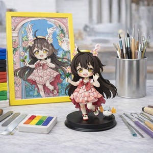 Op de afbeelding: Gedetailleerd beeldje van een cartoonmeisje met lang donker haar, gekleed in een roze jurk en met een bloem in de hand. Een ingelijste foto van hetzelfde meisje staat op de achtergrond. Het beeldje staat op een zwarte voet, met een kleine vogel in de buurt. Diverse kunstbenodigdheden zijn ook zichtbaar.