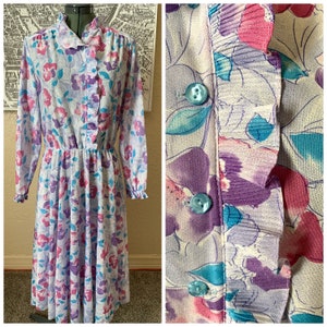 Könnte beinhalten: Ein langärmeliges Kleid mit einem Blumendruck in den Farben Lila, Rosa und Blau. Das Kleid hat einen Rüschenkragen und -ärmel sowie eine eingezogene Taille.