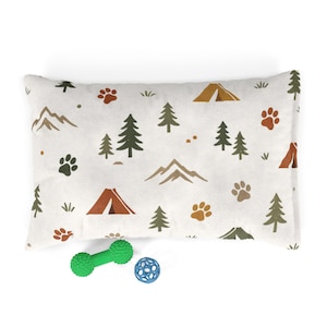 May include: A rectangular pet bed with a white background featuring a camping-themed pattern. The pattern includes brown tents, paw prints, trees, and mountains. A green dumbbell-shaped toy and a blue ball toy are in front of the bed.