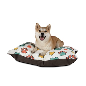 May include: A Shiba Inu dog rests on a square pet bed. The bed has a white top with colorful paw prints, hearts, and leaf designs. The bed's sides are brown.