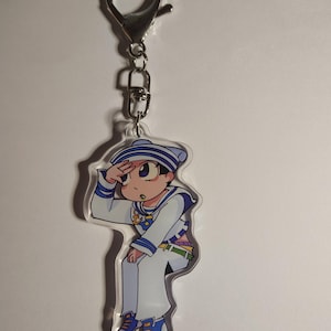 Puede incluir: Un llavero de acrílico transparente con un personaje de dibujos animados que viste un conjunto de marinero blanco y azul con un gorro a juego. El personaje tiene el pelo oscuro, hace un saludo y lleva zapatillas azules. Cierre de mosquetón.