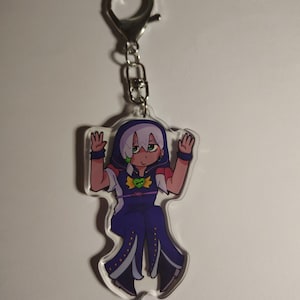Puede incluir: Un llavero de acrílico transparente con un personaje de dibujos animados de cabello violeta claro, que viste un atuendo azul oscuro con capucha, un emblema de corazón verde, detalles amarillos y un cinturón morado, unido a un broche de langosta plateado.