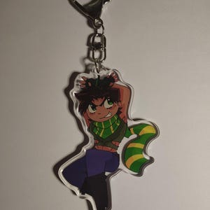 Puede incluir: Un llavero de acrílico transparente con un personaje de dibujos animados de cabello oscuro, ojos verdes y una bufanda a rayas verdes y amarillas. El personaje viste un top corto verde oscuro, pantalones morados y botas negras.