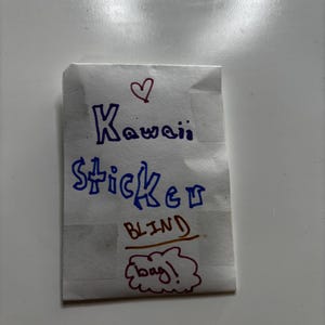 May include: A small, folded paper bag with the words "Kawaii Sticker BLIND bag!" written in blue and red marker. A red heart is drawn above the word "Kawaii". The bag is white with a few vertical lines.