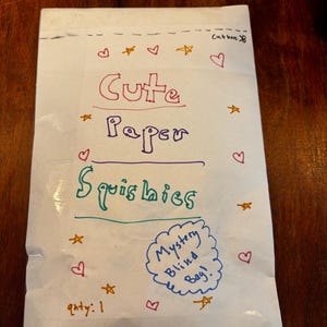 May include: A white paper bag with the words "Cute Paper Squishies" written in colorful marker. The bag is decorated with hearts and stars, and the text "Mystery Blind Bag!" is written in a blue cloud shape. The quantity is marked as 1.