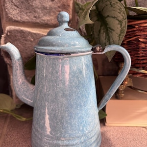 May include: A vintage, light blue enamelware coffee pot with a lid and handle. The pot has a speckled finish and a curved spout. The handle is arched, and the lid has a small knob. The pot is a classic kitchenware item.