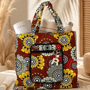 Peut inclure: Un sac fourre-tout avec un motif mandala vibrant dans des tons de jaune, rouge et gris. Le sac possède deux poignées supérieures et une poche frontale zippée avec un porte-clés lettre R doré. L'intérieur contient un livre intitulé CEREAL.