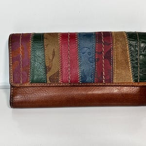 May include: A brown leather wallet with a patchwork design. The patchwork features vertical stripes in various colors, including purple, green, red, blue, and tan. The wallet has a flap closure and a brown leather base.