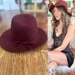 May include: A burgundy-colored hat with a wide brim and a small bow detail. The hat has a rounded crown and is made of a soft material. The hat is a stylish accessory.