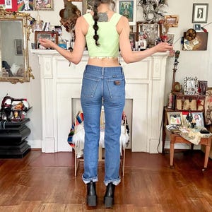 Puede incluir: Un top corto verde claro con cuello cuadrado y ribete floral, combinado con vaqueros de mezclilla azules y botas de plataforma negras. Una gorra de b&eacute;isbol negra se lleva al rev&eacute;s. El conjunto se exhibe en una habitaci&oacute;n con chimenea blanca y varios objetos decorativos.