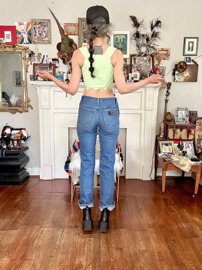 Puede incluir: Un top corto verde claro con cuello cuadrado y ribete floral, combinado con vaqueros de mezclilla azules y botas de plataforma negras. Una gorra de b&eacute;isbol negra se lleva al rev&eacute;s. El conjunto se exhibe en una habitaci&oacute;n con chimenea blanca y varios objetos decorativos.