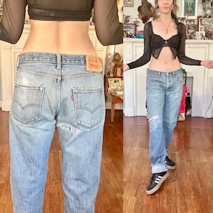 May include: Light blue denim jeans with distressed details, featuring a classic five-pocket design and a red Levi's label. The jeans have a straight leg fit and are worn with a black top and sneakers.