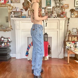 Puede incluir: Vaqueros de mezclilla azul claro con el logo de Wrangler en el bolsillo trasero. Los vaqueros se combinan con un top corto estampado y botas de plataforma negras. El atuendo se captura en una toma de cuerpo entero, mostrando un estilo casual de inspiraci&oacute;n vintage.