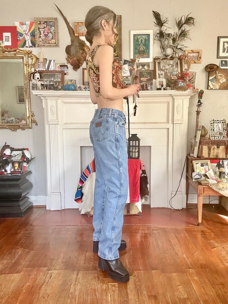 Puede incluir: Vaqueros de mezclilla azul claro con el logo de Wrangler en el bolsillo trasero. Los vaqueros se combinan con un top corto estampado y botas de plataforma negras. El atuendo se captura en una toma de cuerpo entero, mostrando un estilo casual de inspiraci&oacute;n vintage.