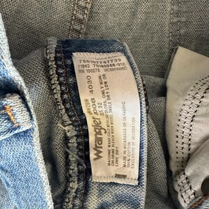 Puede incluir: Primer plano de unos vaqueros de mezclilla azul claro con una etiqueta Wrangler. La etiqueta muestra la marca, la talla 32x32 y las instrucciones de cuidado. Los vaqueros son 100% algod&oacute;n y tienen un dise&ntilde;o cl&aacute;sico.