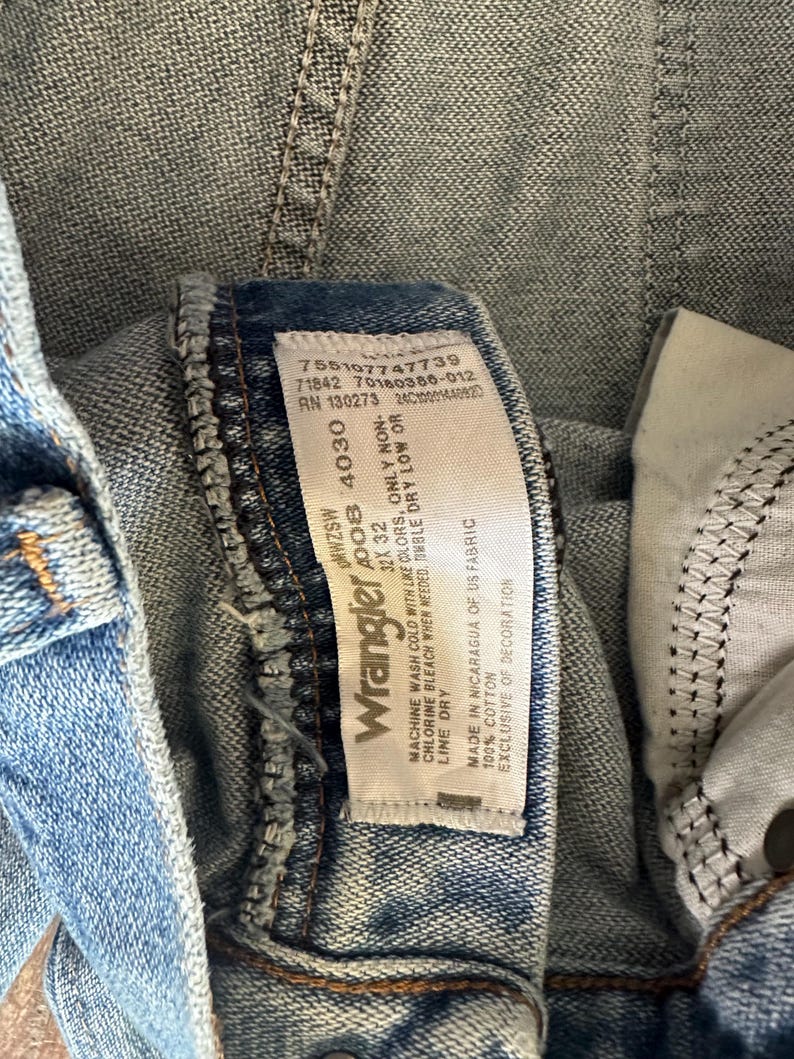 Puede incluir: Primer plano de unos vaqueros de mezclilla azul claro con una etiqueta Wrangler. La etiqueta muestra la marca, la talla 32x32 y las instrucciones de cuidado. Los vaqueros son 100% algod&oacute;n y tienen un dise&ntilde;o cl&aacute;sico.