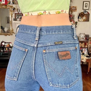 Puede incluir: Vaqueros Wrangler de mezclilla azul claro con un corte cl&aacute;sico. Los vaqueros presentan un parche de cuero marr&oacute;n con el logo de Wrangler en el bolsillo trasero. Los bolsillos traseros tienen un dise&ntilde;o de costura caracter&iacute;stico. Los vaqueros se llevan con un top corto verde claro.