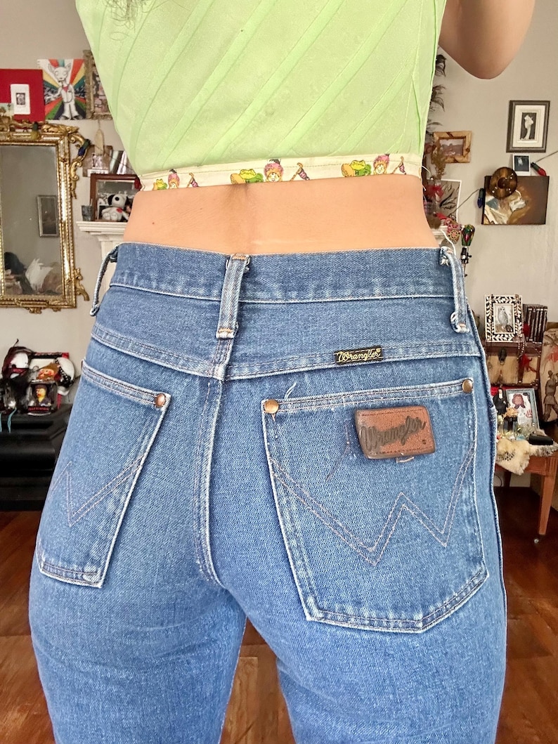 Puede incluir: Vaqueros Wrangler de mezclilla azul claro con un corte cl&aacute;sico. Los vaqueros presentan un parche de cuero marr&oacute;n con el logo de Wrangler en el bolsillo trasero. Los bolsillos traseros tienen un dise&ntilde;o de costura caracter&iacute;stico. Los vaqueros se llevan con un top corto verde claro.