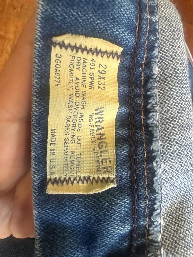 Puede incluir: Primer plano de una etiqueta de vaqueros de mezclilla azul. La etiqueta dice "WRANGLER NO-FAULT DENIM" con la talla 29x32. La etiqueta tambi&eacute;n incluye instrucciones de lavado y "MADE IN U.S.A."
