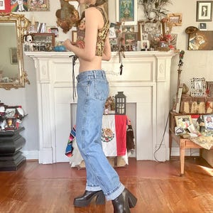 Puede incluir: Una mujer con un top corto estampado, vaqueros azul claro y botas de plataforma negras. El top tiene un dise&ntilde;o floral dorado y marr&oacute;n. Los vaqueros est&aacute;n doblados en los tobillos. Las botas tienen un tac&oacute;n grueso. El fondo incluye una chimenea y cuadros.