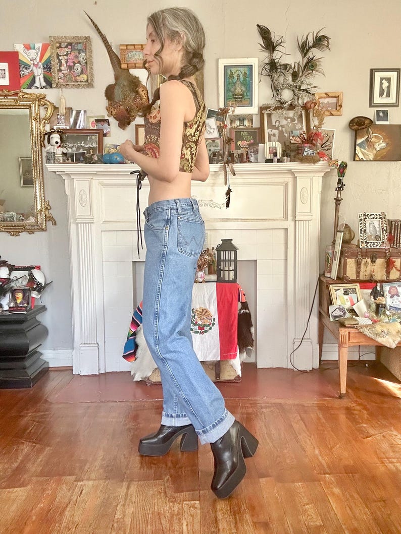 Puede incluir: Una mujer con un top corto estampado, vaqueros azul claro y botas de plataforma negras. El top tiene un dise&ntilde;o floral dorado y marr&oacute;n. Los vaqueros est&aacute;n doblados en los tobillos. Las botas tienen un tac&oacute;n grueso. El fondo incluye una chimenea y cuadros.
