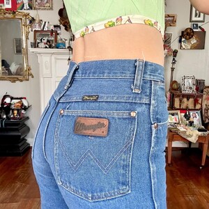 Puede incluir: Vaqueros Wrangler de mezclilla azul claro con un parche de cuero marr&oacute;n en el bolsillo trasero. Los vaqueros tienen una cintura alta y un dise&ntilde;o cl&aacute;sico de cinco bolsillos. El modelo lleva un top corto verde claro.