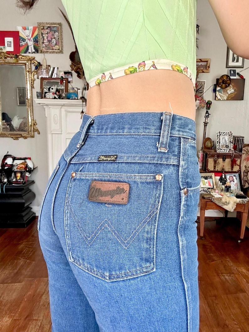 Puede incluir: Vaqueros Wrangler de mezclilla azul claro con un parche de cuero marr&oacute;n en el bolsillo trasero. Los vaqueros tienen una cintura alta y un dise&ntilde;o cl&aacute;sico de cinco bolsillos. El modelo lleva un top corto verde claro.
