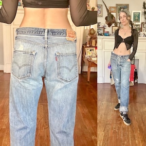 May include: Light blue denim jeans with a faded wash and distressed details. The jeans feature classic five-pocket styling, a button and zipper closure, and a red logo patch on the back. The jeans are cuffed at the ankles.