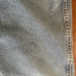 Puede incluir: Primer plano de tejido vaquero azul claro. El denim tiene un aspecto texturizado con detalles de tejido visibles. Una costura recorre verticalmente el lado derecho, con una puntada en contraste. El tono general es descolorido y desgastado.